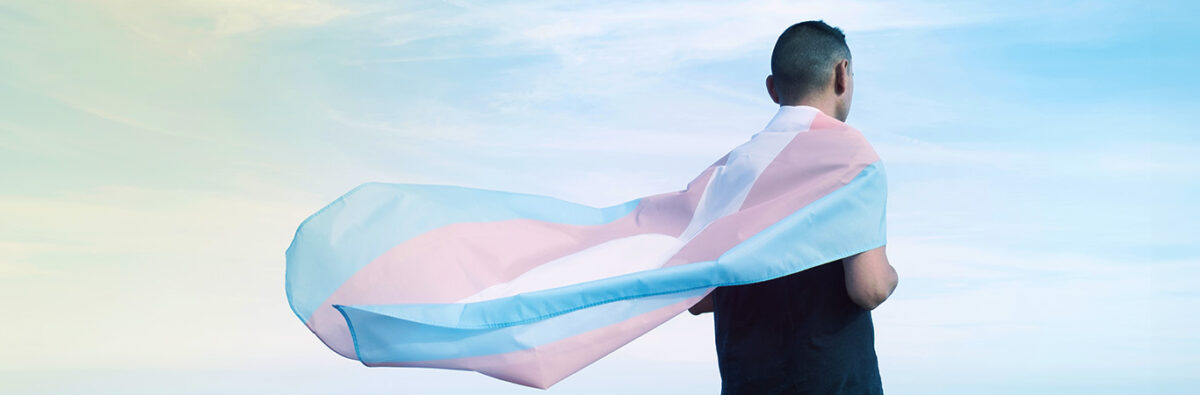Trans man in black with trans flagged wrapped around, facing sky horizon