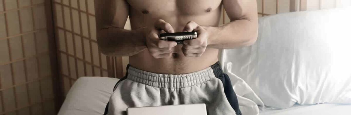 Muscular man doing some research on his phone and his laptop wearing track pants and shirtless