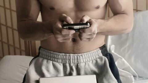Muscular man doing some research on his phone and his laptop wearing track pants and shirtless