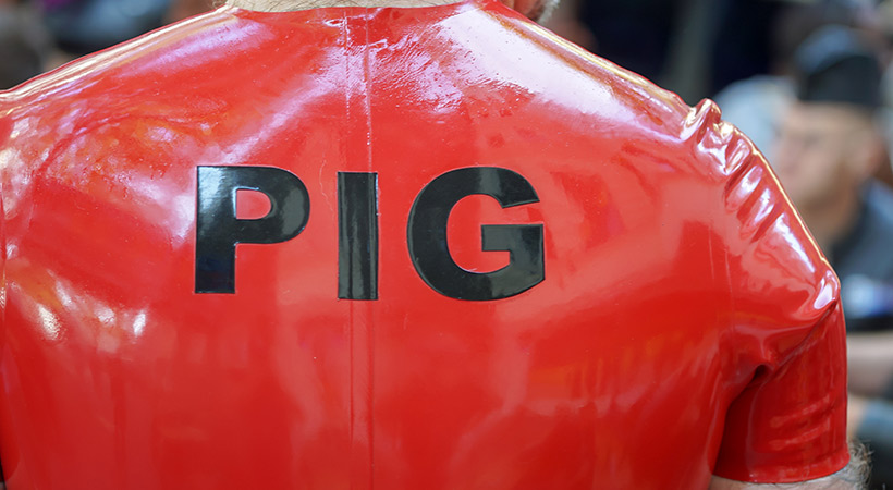 man wears red rubber fetish wear with the word PIG in black letters on the back