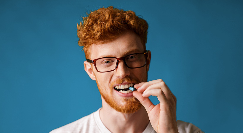 red haired man in glasses biting prep pill on blue background