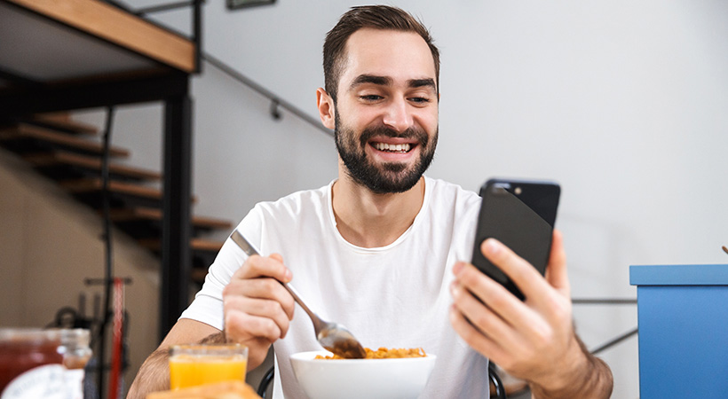 man eats bowl of cereal while smiling at a message on his mobile phone