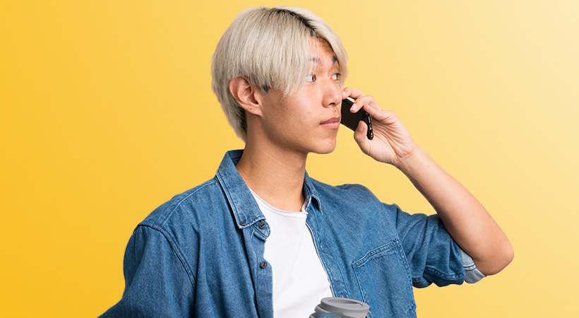 young asian man with blonde hair holds coffee cup while on phone