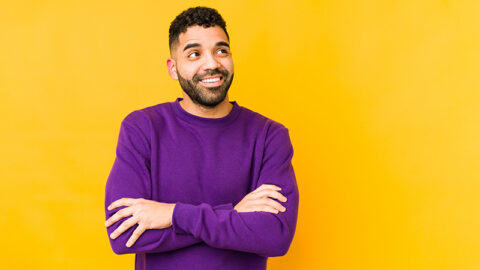 stubbled man wears purple sweater with arms folded against yellow background