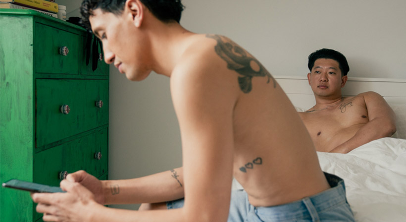 Shirtless man on phone at end of bed, partner in bed under covers looking jealous