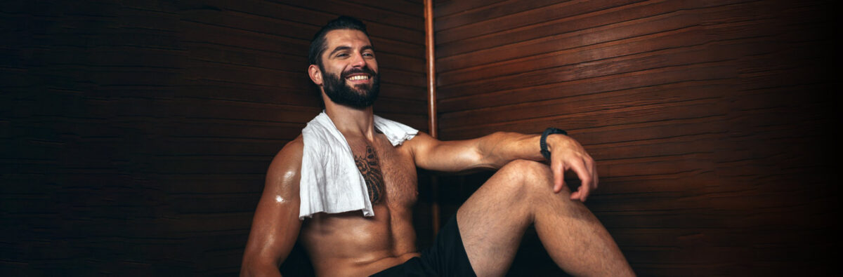 tattood man sits in a gay sauna with towel round his neck smiling