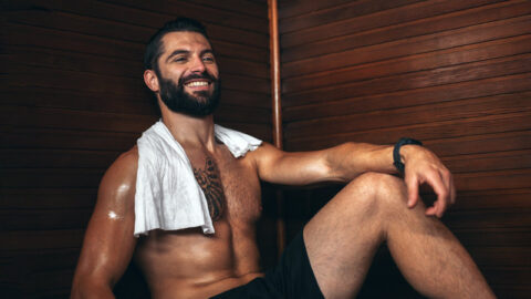 tattood man sits in a gay sauna with towel round his neck smiling