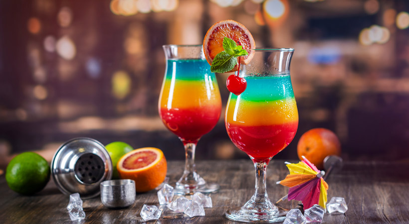 two rainbow cocktails with shaker and fruit garnishes