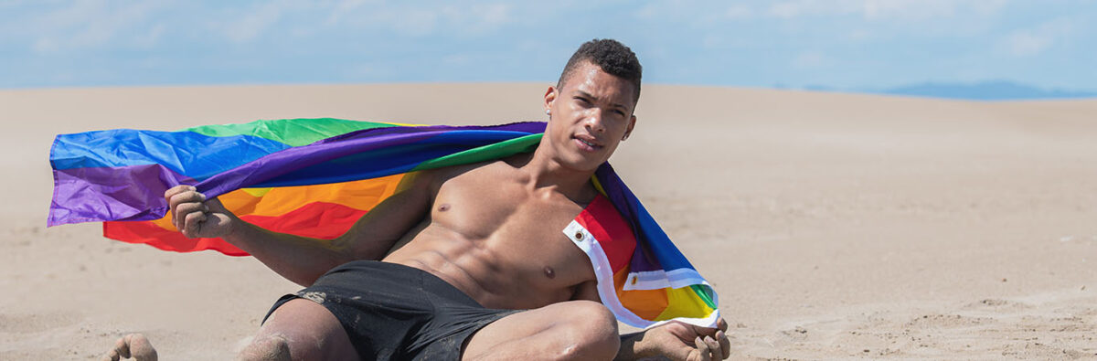 young black guy lays down on beach wrapped in pride flag hero