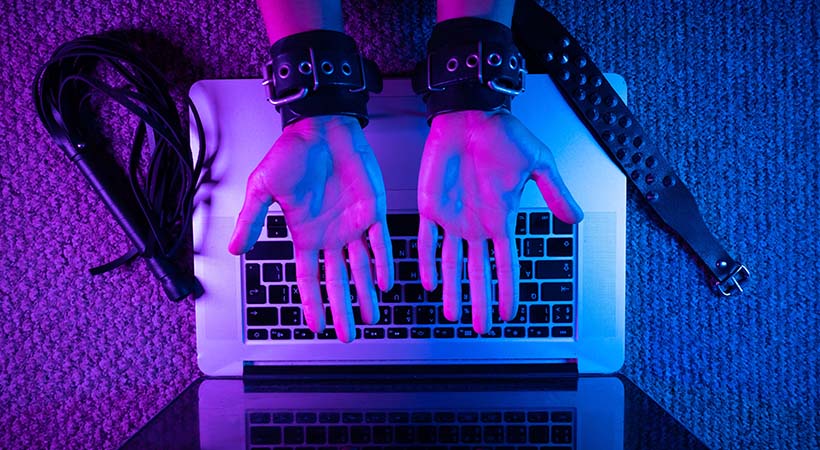 hands in bdsm handcuffs with laptop in neon light on dark background