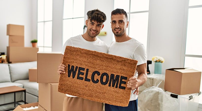 Two hispanic men couple hugging each other holding welcome doormat at new home