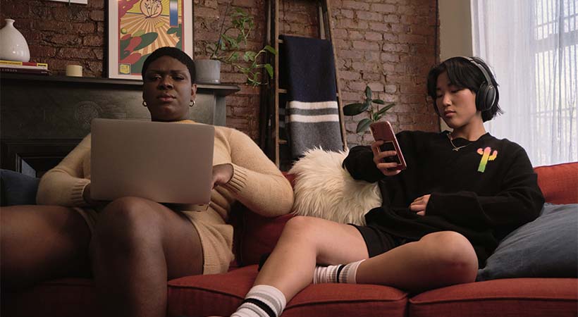 Two roommates on a couch using laptop and phone