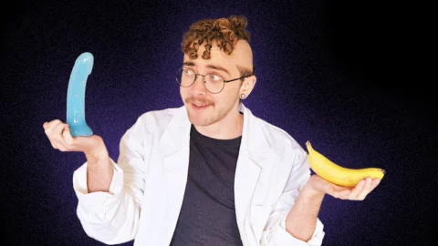 White transmasc person in white coat holding banana and dildo on purple black background