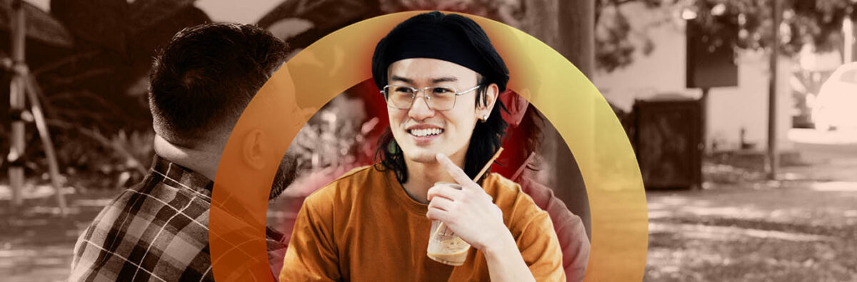 Vietnamese man in orange shirt holding iced coffee, smiling at friend in a park